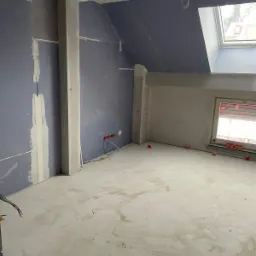 Poddasze w trakcie adaptacji: szare płyty na skosach dachu, częściowo zaszpachlowane łączenia, widoczne instalacje elektryczne i okno dachowe.