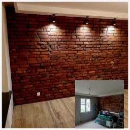 Ceglana ściana w salonie z trzema czarnymi reflektorami punktowymi, widok na podłogę laminowaną i fragment drugiego pomieszczenia w trakcie prac wykończeniowych z oknem i stosem materiałów...