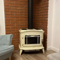 Elegancki, kremowy piec wolnostojący z czarnym kominem na tle ściany z czerwonej cegły dekoracyjnej, uzupełniony jasnym panelem podłogowym i fragmentami mebli w stonowanych kolorach.