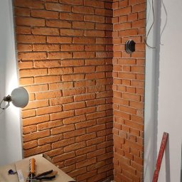SOLID HOME remonty i wykończenia wnętrz - Fragment ściany wykończonej dekoracyjną cegłą w odcieniach czerwieni, widoczne narzędzia i materiały budowlane w trakcie prac wykończeniowych, lampka oświetlająca fragment ściany, otwór w ścianie...