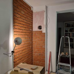SOLID HOME remonty i wykończenia wnętrz - Fragment ściany z cegły klinkierowej w trakcie montażu, oświetlony lampą warsztatową, widoczny fragment drabiny i narzędzia na stole, otwór w ścianie przygotowany pod instalację.