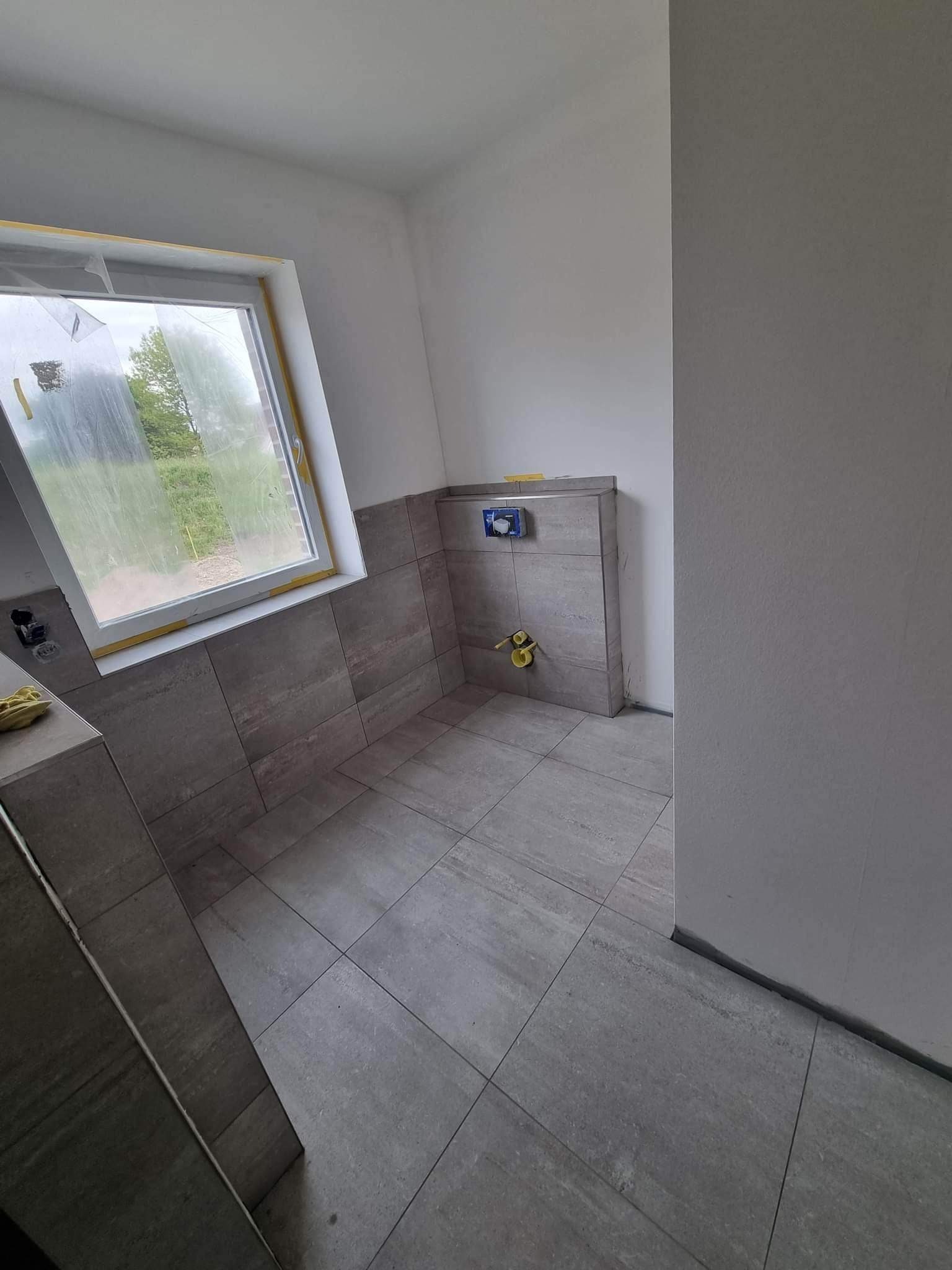 Wnętrze łazienki w trakcie prac wykończeniowych; widoczne szare płytki na podłodze i ścianach, zabudowa podtynkowego WC, zabezpieczone folią okno dachowe z widokiem na zieleń na zewnątrz.