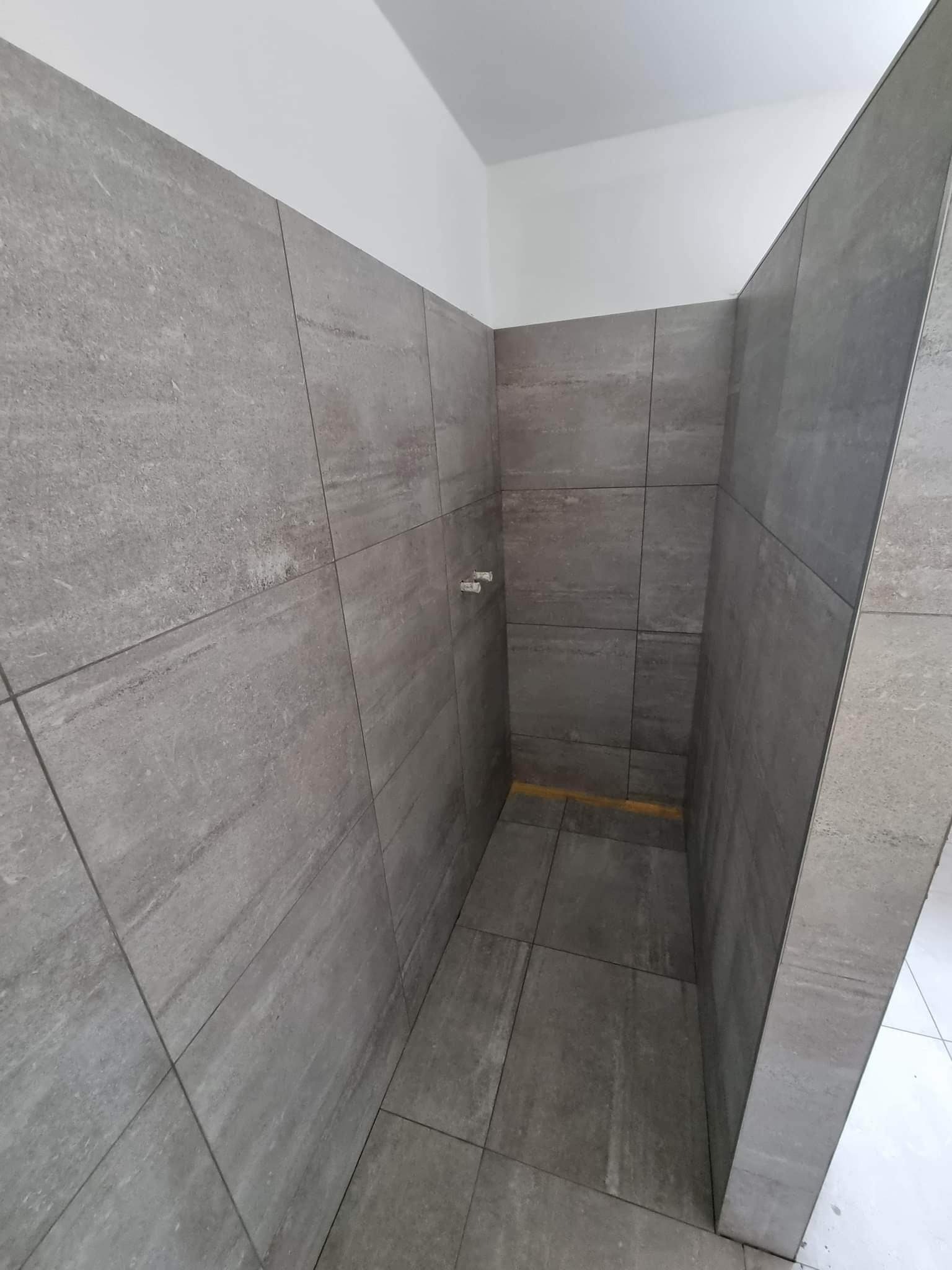 Świeżo wykafelkowana łazienka z szarymi, matowymi płytkami imitującymi kamień, widoczny fragment instalacji wodnej na ścianie i żółta taśma zabezpieczająca na podłodze.