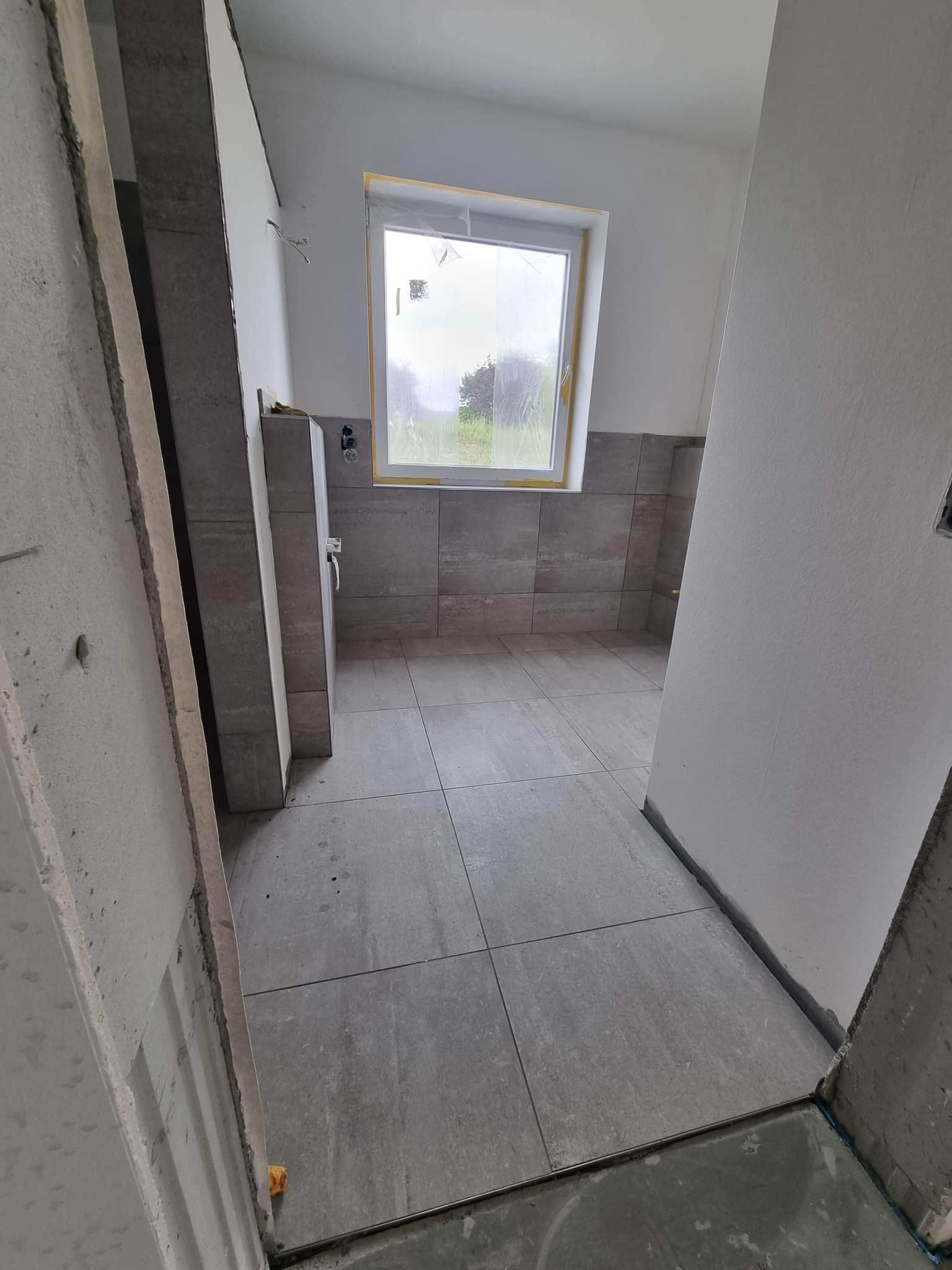 Wnętrze łazienki w trakcie remontu: szare płytki na podłodze i częściowo na ścianach, okno z widokiem na zieleń, surowe ściany w trakcie wykończenia.