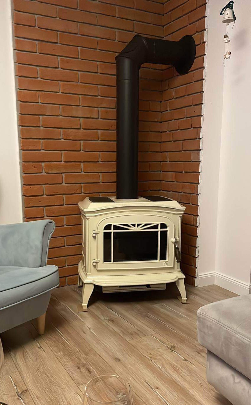 Elegancki, kremowy piec wolnostojący z czarnym kominem na tle ściany z czerwonej cegły dekoracyjnej, uzupełniony jasnym panelem podłogowym i fragmentami mebli w stonowanych kolorach.