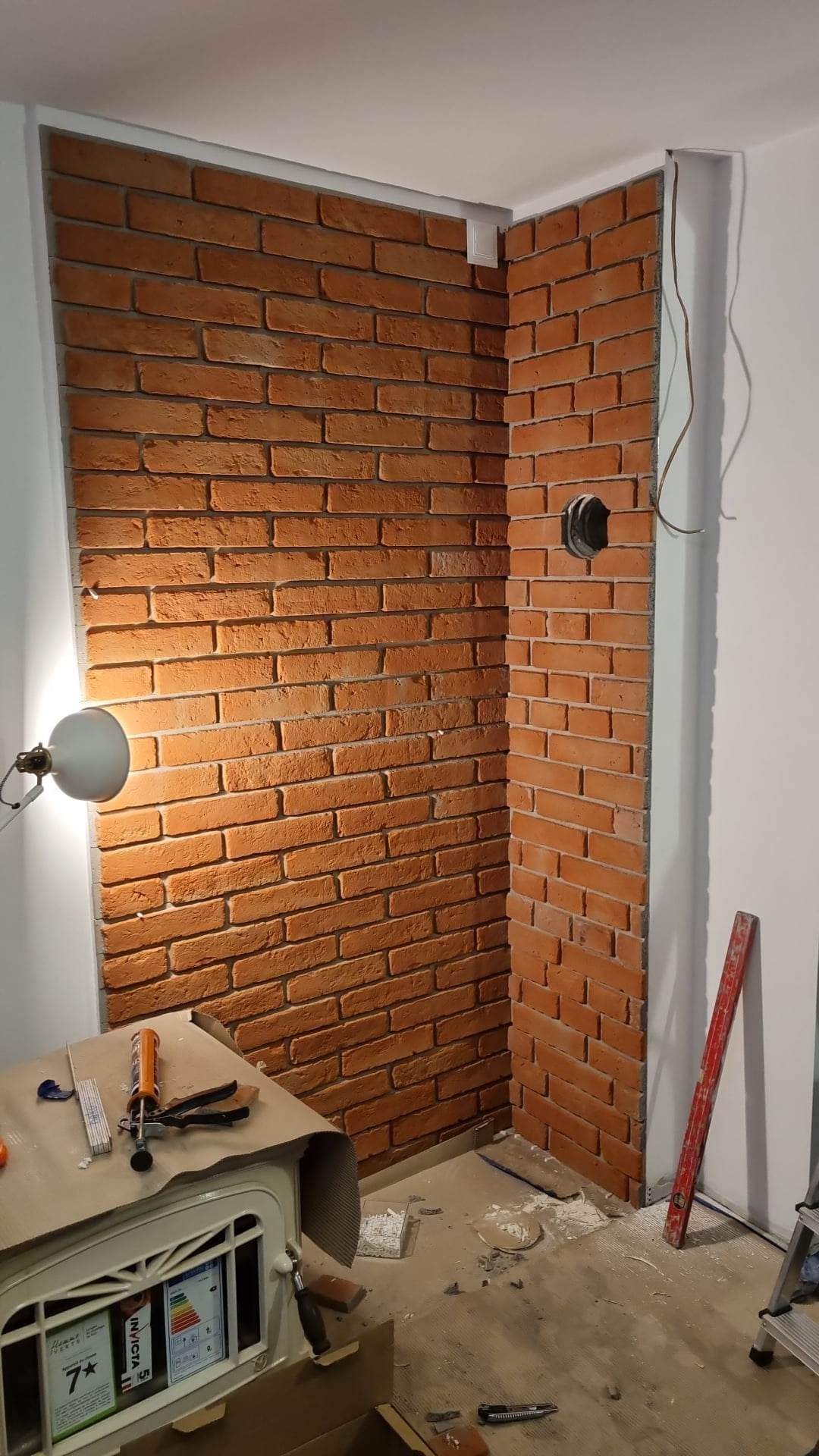 Fragment ściany wykończonej dekoracyjną cegłą w odcieniach czerwieni, widoczne narzędzia i materiały budowlane w trakcie prac wykończeniowych, lampka oświetlająca fragment ściany, otwór w ścianie...