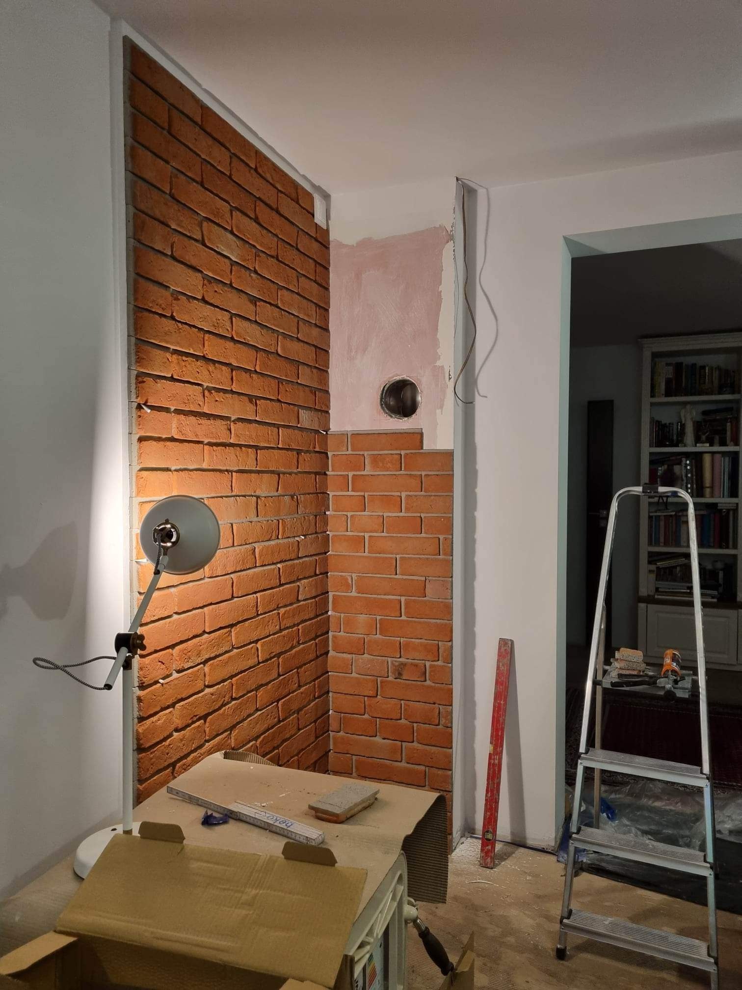 Fragment ściany z cegły klinkierowej w trakcie montażu, oświetlony lampą warsztatową, widoczny fragment drabiny i narzędzia na stole, otwór w ścianie przygotowany pod instalację.