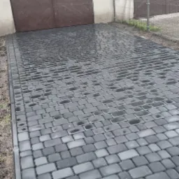 Świeżo ułożona, mokra, szara kostka brukowa prowadząca do brązowych, dwuskrzydłowych drzwi garażowych. Widoczne obrzeża i częściowo metalowe ogrodzenie.