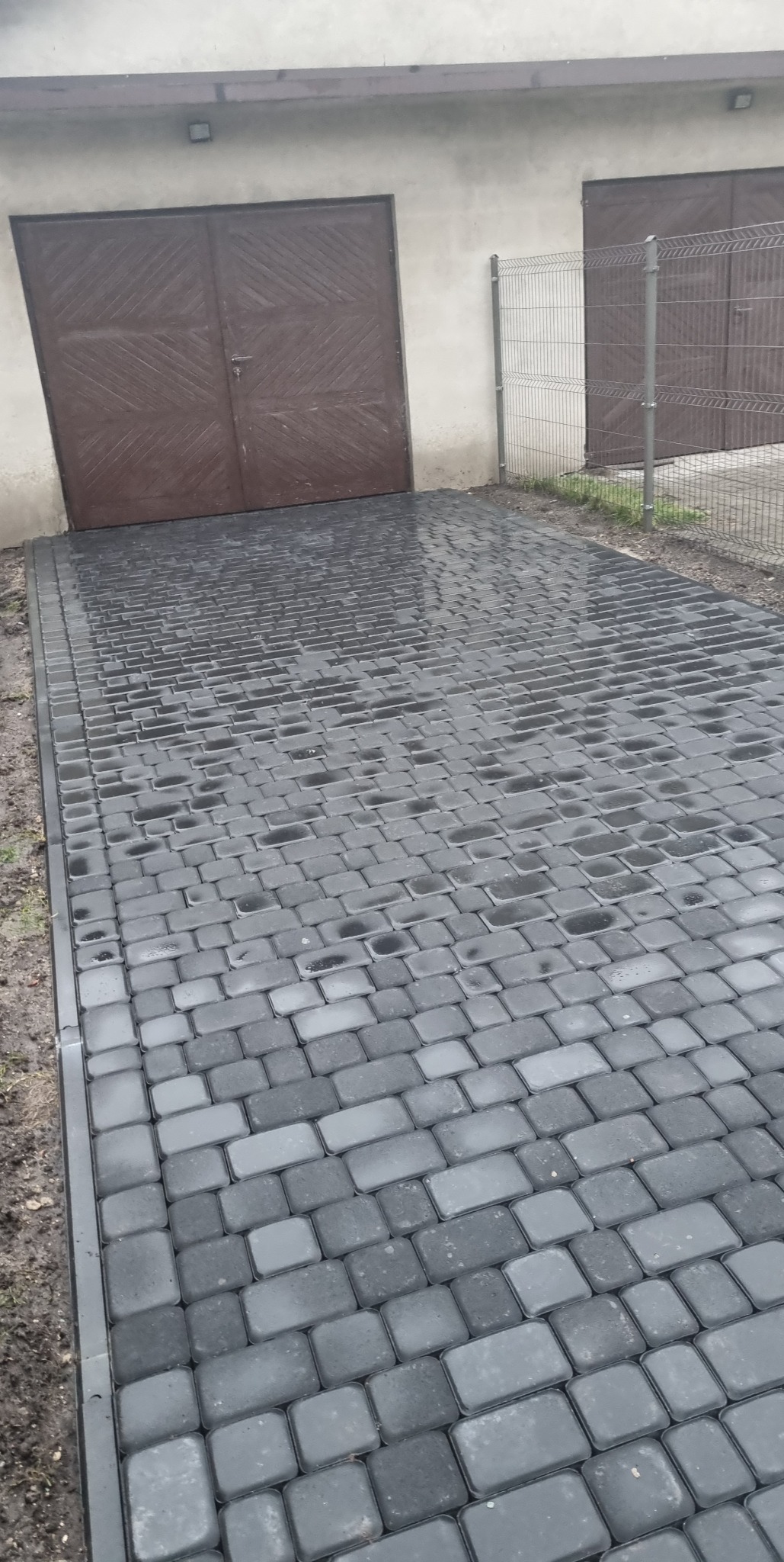 Świeżo ułożona, mokra, szara kostka brukowa prowadząca do brązowych, dwuskrzydłowych drzwi garażowych. Widoczne obrzeża i częściowo metalowe ogrodzenie.