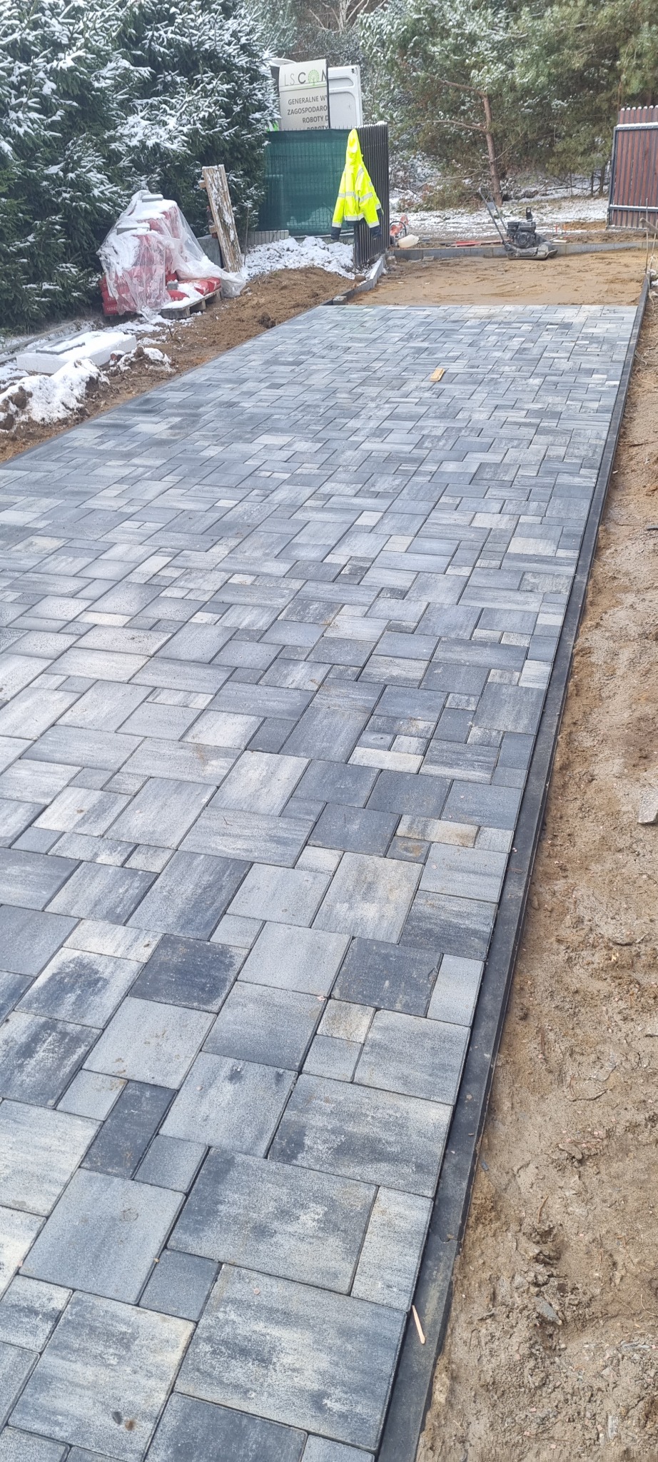 Ułożona szara kostka brukowa o nieregularnym wzorze, widoczny fragment podjazdu z podsypką piaskową po bokach, w tle maszyna do ubijania gruntu, żółta kurtka robocza i częściowo ośnieżone drzewa.