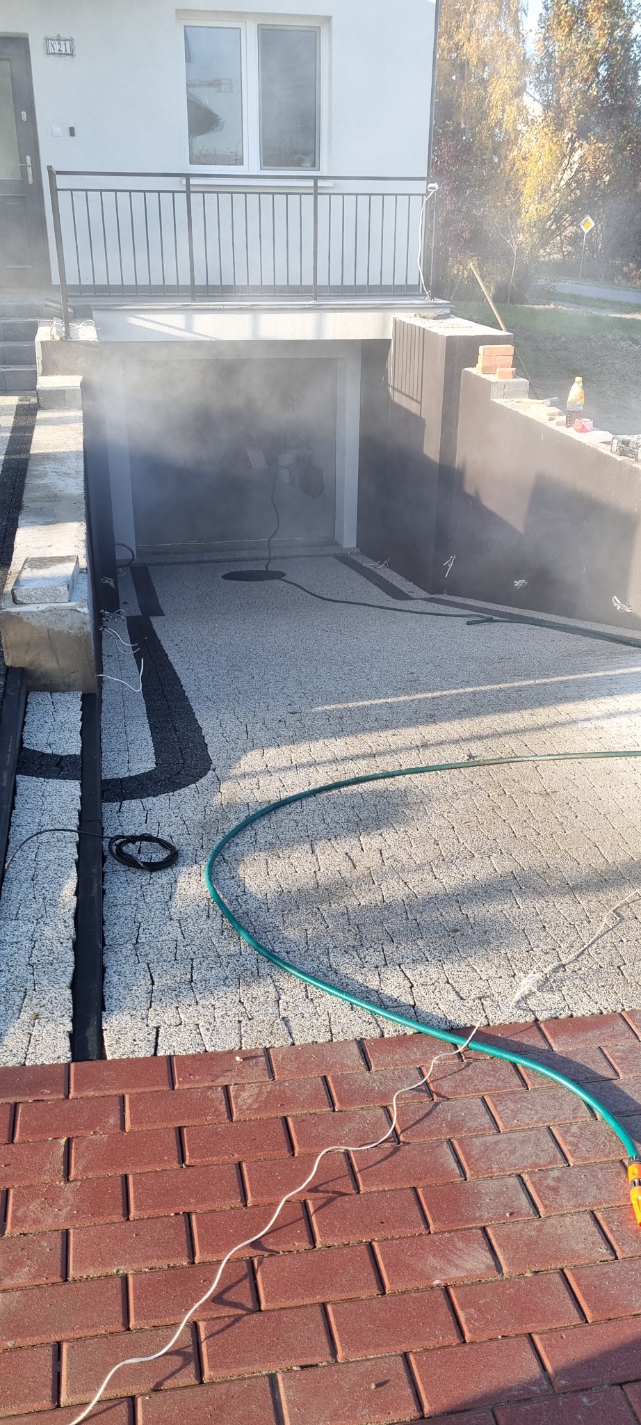 Nowa, jasnoszara nawierzchnia z kostki brukowej prowadząca do garażu, z widocznymi czarnymi fugami i zielonym wężem ogrodowym. W tle widać zamglenie.