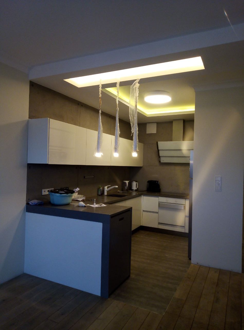 Nowoczesna kuchnia z białymi szafkami, szarą ścianą i blatem, oświetlona przez podwieszane lampy w folii ochronnej oraz wbudowane światła w suficie.