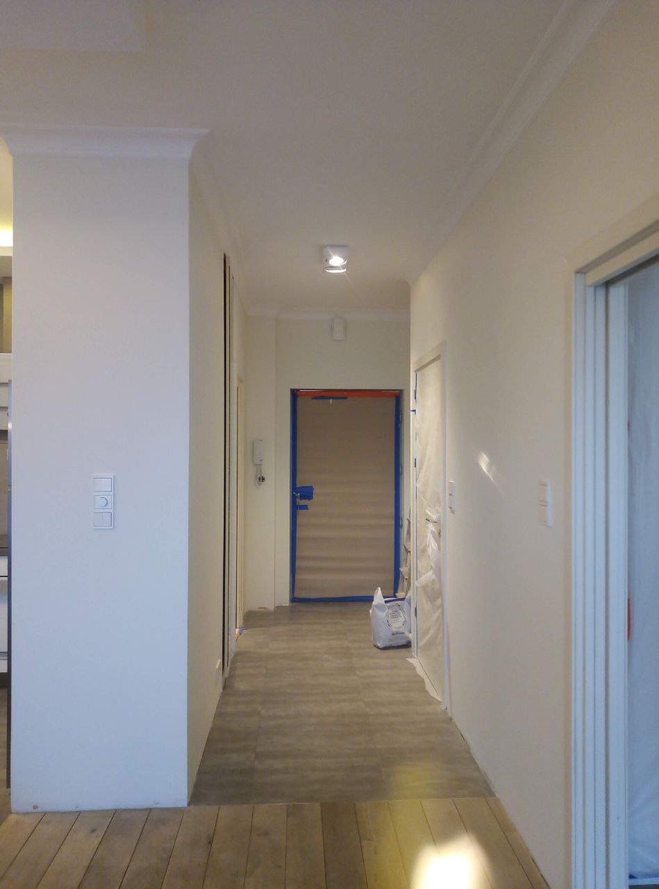 Korytarz w trakcie prac wykończeniowych; ściany pomalowane na biało, drzwi zabezpieczone folią malarską, widoczna listwa dekoracyjna pod sufitem, podłoga częściowo zakryta.