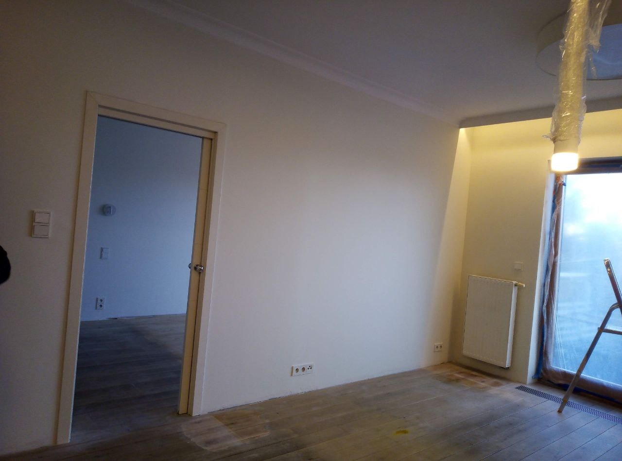 Wnętrze pokoju po odświeżeniu, z pomalowanymi ścianami na jasny kolor, widoczne drewniane podłogi, grzejnik i drabina przy oknie, lampa sufitowa zabezpieczona folią.