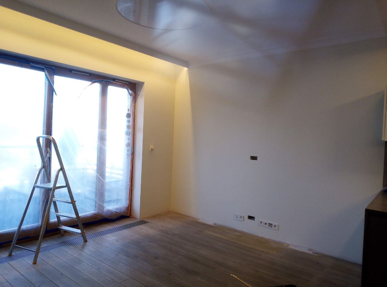 Pokój w trakcie przygotowań do malowania: okno zabezpieczone folią, drabina przystawiona, ściany zagruntowane, widoczne gniazdka elektryczne.