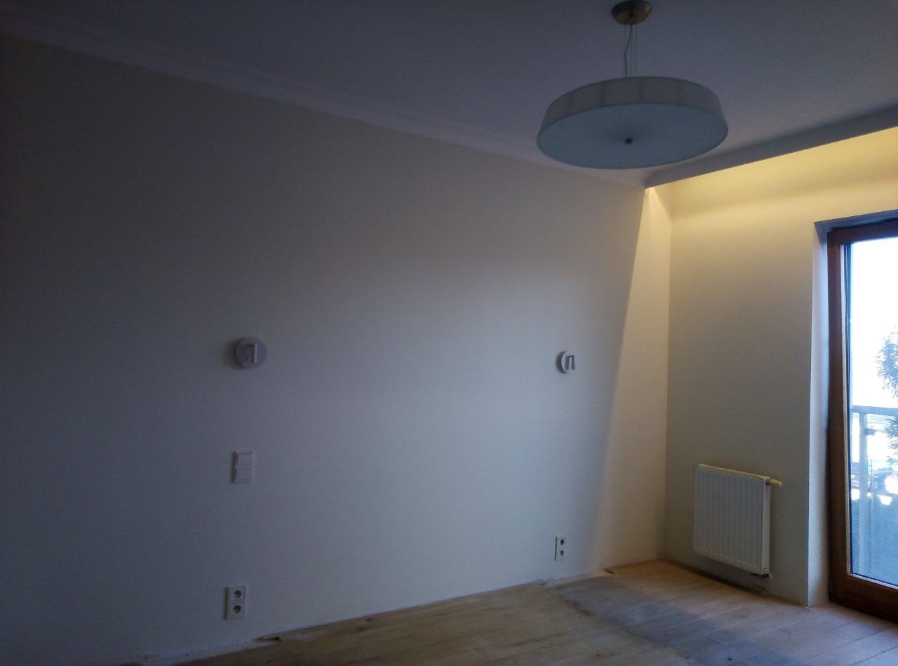 Jasne wnętrze pokoju po malowaniu ścian na biało, widoczne gniazdka elektryczne i czujniki na ścianie, drewniana podłoga i okno z widokiem na zewnątrz.