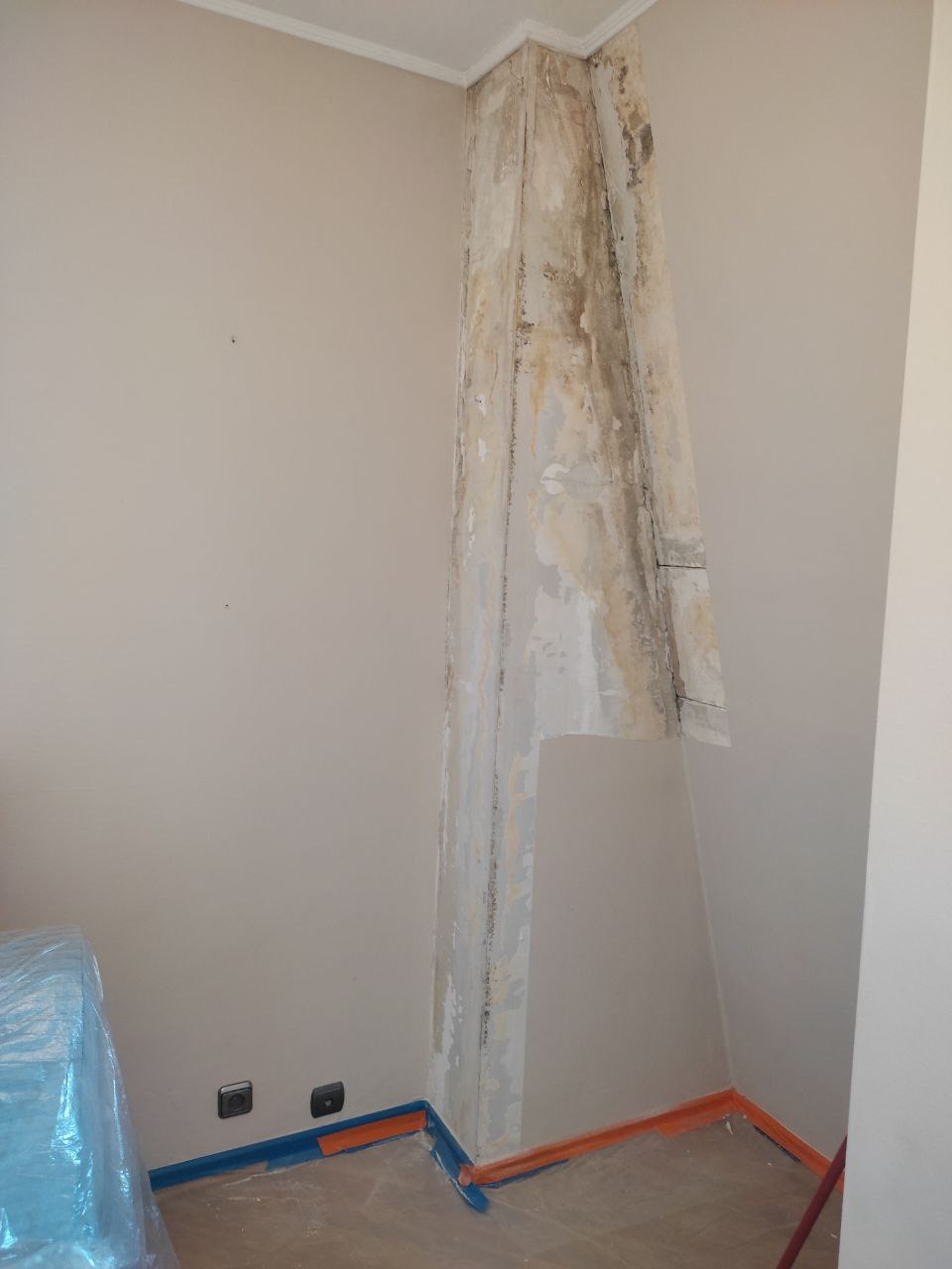 Pokój w trakcie przygotowania do odświeżenia: zabezpieczone listwy przypodłogowe taśmą malarską, ściany częściowo zdarta stara farba, widoczne plamy i uszkodzenia na filarze narożnym.