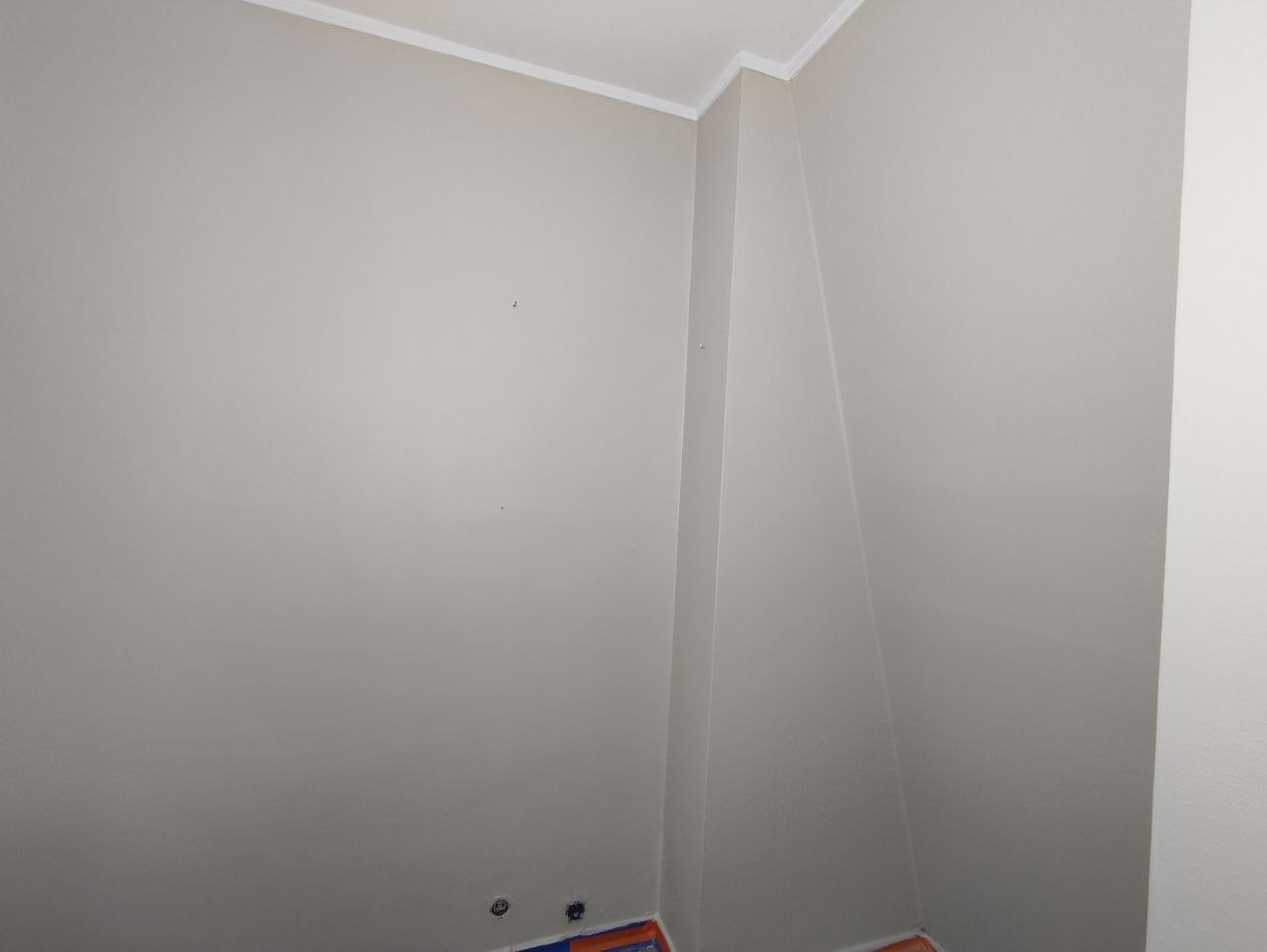 Świeżo pomalowane, jasnoszare ściany w rogu pomieszczenia, z widocznymi otworami po gniazdkach elektrycznych zabezpieczonych taśmą malarską przy podłodze oraz białą listwą przypodłogową.