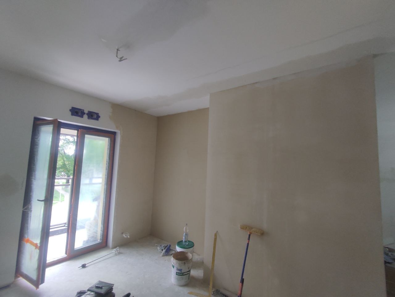 Wnętrze w trakcie prac przygotowawczych do malowania: ściany pokryte gładzią, widoczne narzędzia malarskie, otwarte okno balkonowe z widokiem na zieleń.