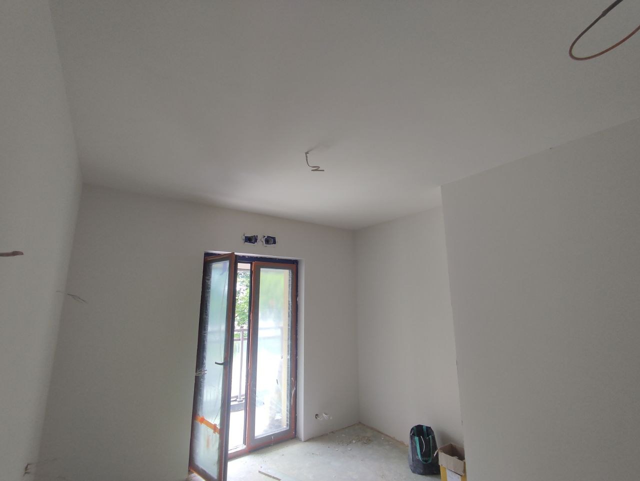 Wnętrze pokoju po świeżym pomalowaniu na biało, widoczne ślady po pracach wykończeniowych, otwarte okno balkonowe i wystające kable ze ściany.