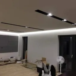 Wnętrze pokoju w trakcie prac wykończeniowych: widoczne kartony, wentylator oraz zamontowane oświetlenie sufitowe LED i panele ścienne.