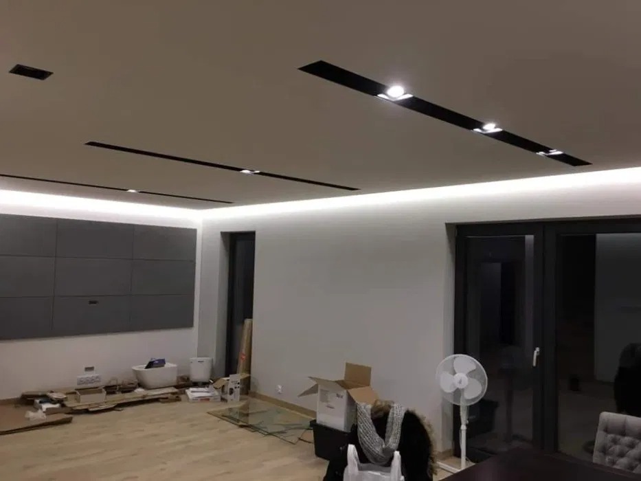 Wnętrze pokoju w trakcie prac wykończeniowych: widoczne kartony, wentylator oraz zamontowane oświetlenie sufitowe LED i panele ścienne.