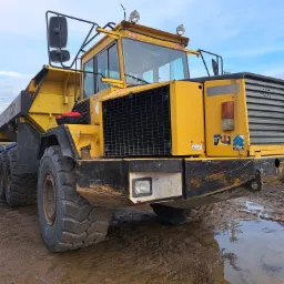 Żółta wywrotka przegubowa Volvo BM na błotnistym placu budowy, widok z przodu na mokre opony i detale kabiny, w tle niebieskie niebo i rury.