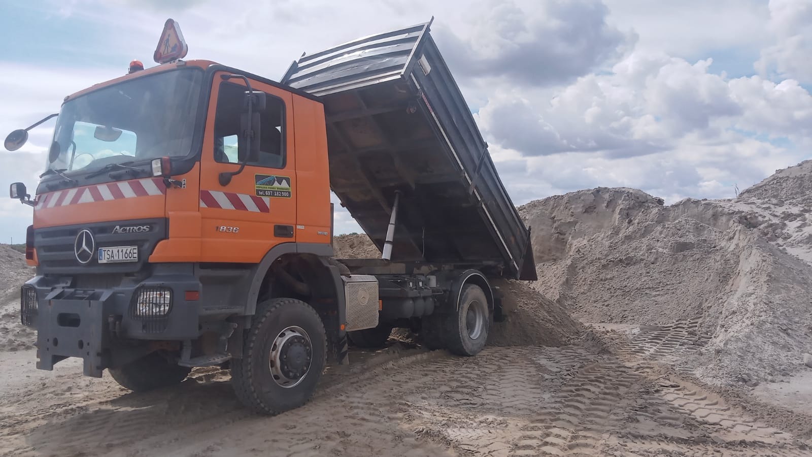 Pomarańczowa wywrotka Mercedes Actros B36 wysypuje piasek na placu budowy, widoczne ślady opon na piasku i chmurne niebo w tle.