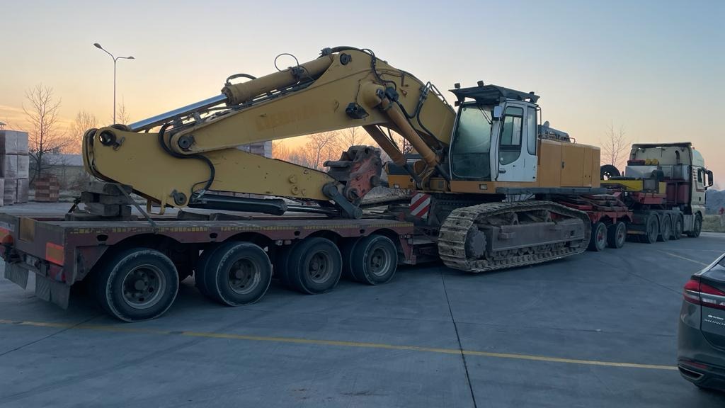Żółta koparka gąsienicowa marki Liebherr transportowana na niskopodwoziowej naczepie ciągniętej przez ciężarówkę, widok z boku na betonowym placu o zmierzchu.