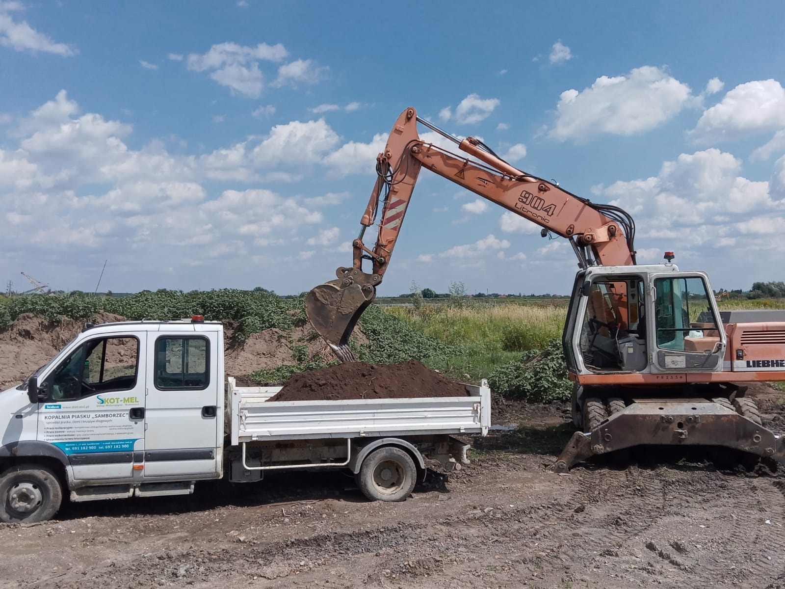 Pomarańczowa koparka Liebherr 904 Litronic nabiera ziemię do białej ciężarówki firmy SKOT-MEL na placu budowy w słoneczny dzień.