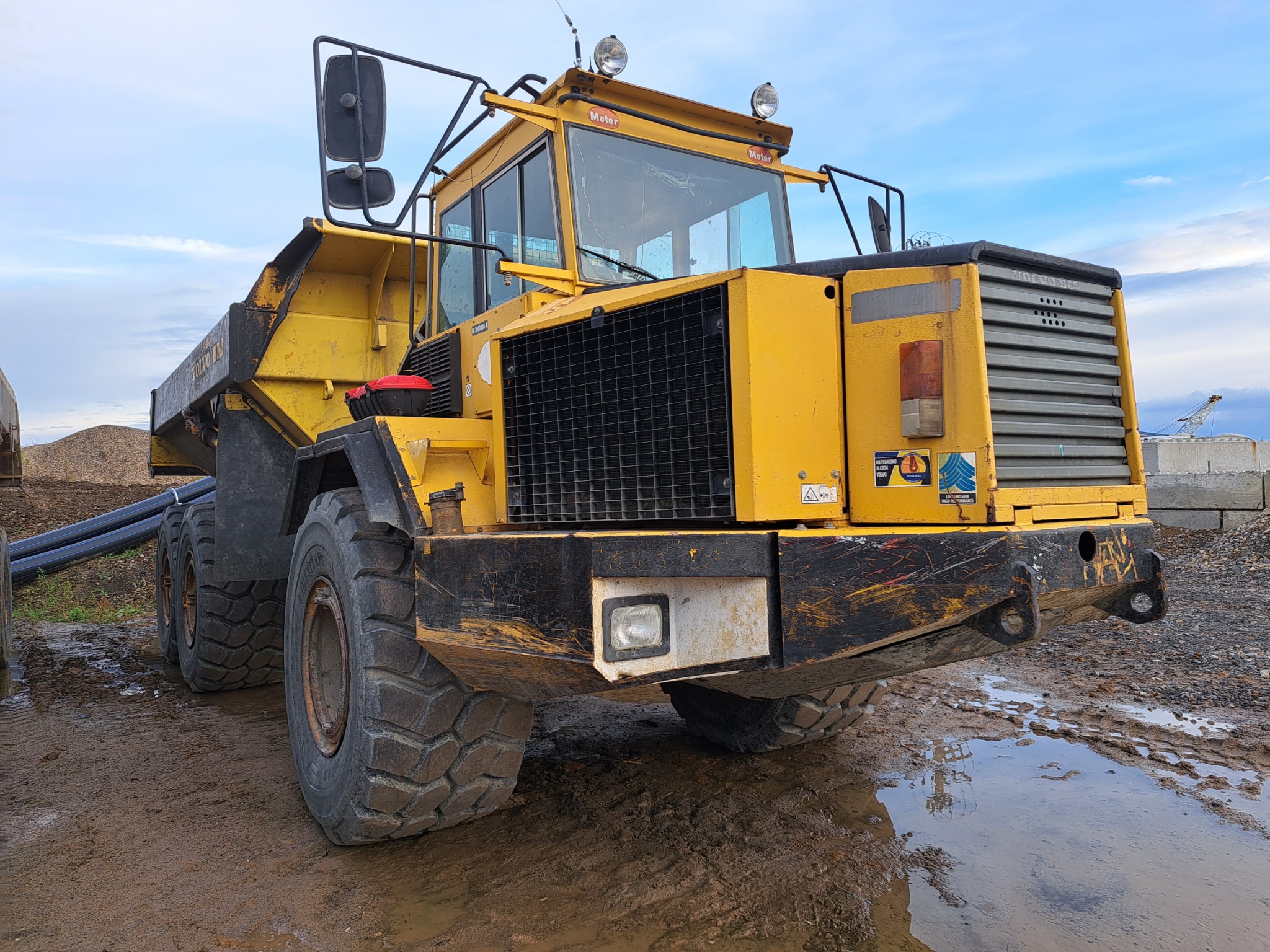 Żółta wywrotka przegubowa Volvo BM na błotnistym placu budowy, widok z przodu na mokre opony i detale kabiny, w tle niebieskie niebo i rury.