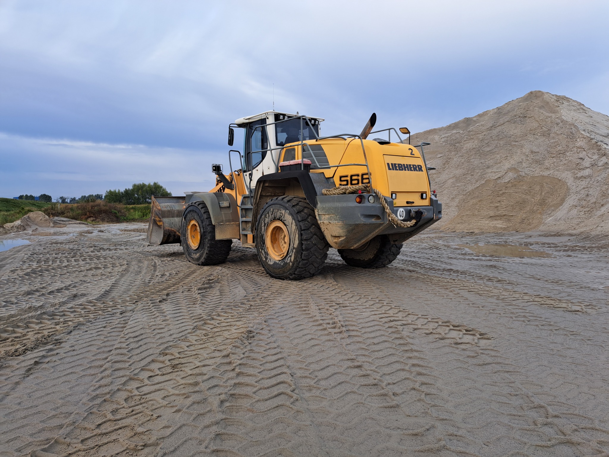 Żółta ładowarka kołowa Liebherr 566 stoi na piaszczystym terenie z widocznymi śladami opon, w tle hałda piasku i pochmurne niebo.