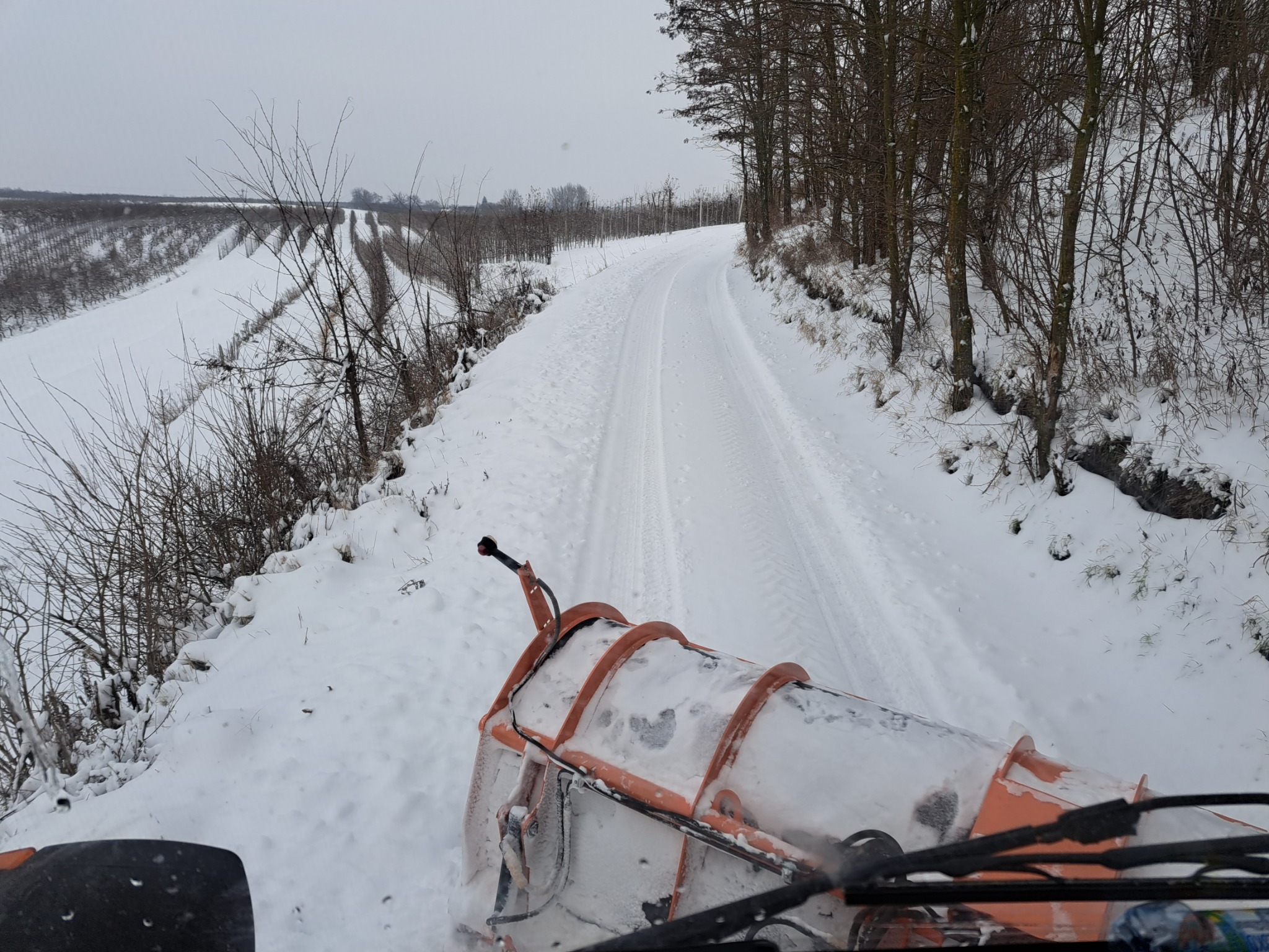 Pomarańczowy pług odśnieżający toruje drogę przez zasypany śniegiem obszar wiejski, widok z kabiny maszyny na zaśnieżoną ścieżkę między polami.