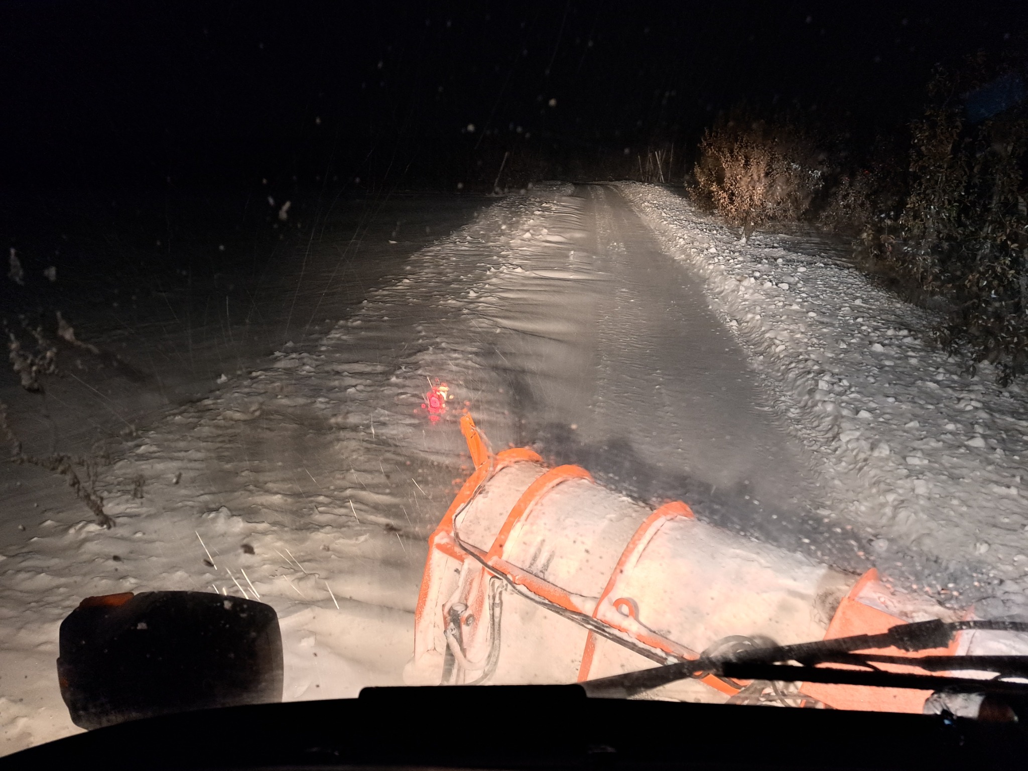 Widok z kabiny pojazdu na odśnieżarkę wirnikową w trakcie pracy na ośnieżonej drodze nocą, widoczne drobinki śniegu na szybie.