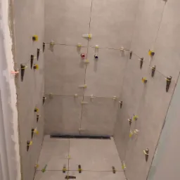 Wnętrze kabiny prysznicowej w trakcie układania jasnych, kwadratowych płytek. Widoczne systemy poziomowania płytek: żółte klipsy i czarne kliny, oraz rury instalacji wodnej.