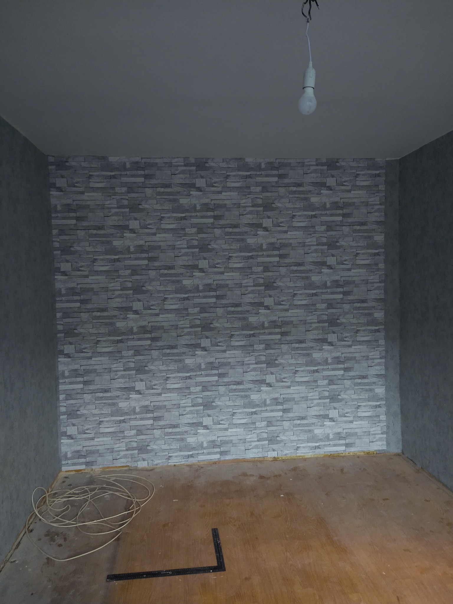 Pokój w trakcie tapetowania, ściana pokryta tapetą imitującą szarą cegłę, kabel elektryczny zwisający z sufitu, na podłodze leżą zwinięte kable i kątownik.
