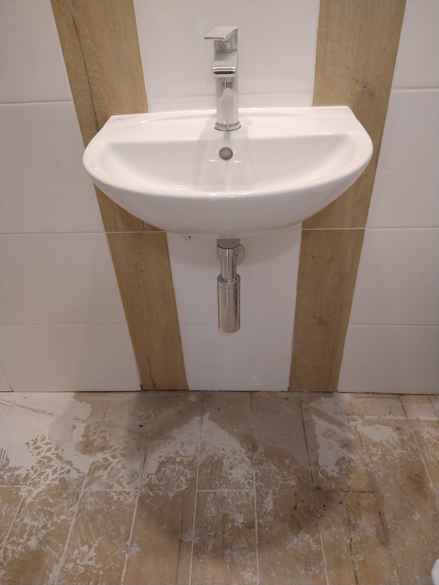 Umywalka wisząca na ścianie wyłożonej białymi płytkami z wstawkami imitującymi drewno. Podłoga zabrudzona po remoncie, widoczne ślady po zaprawie.