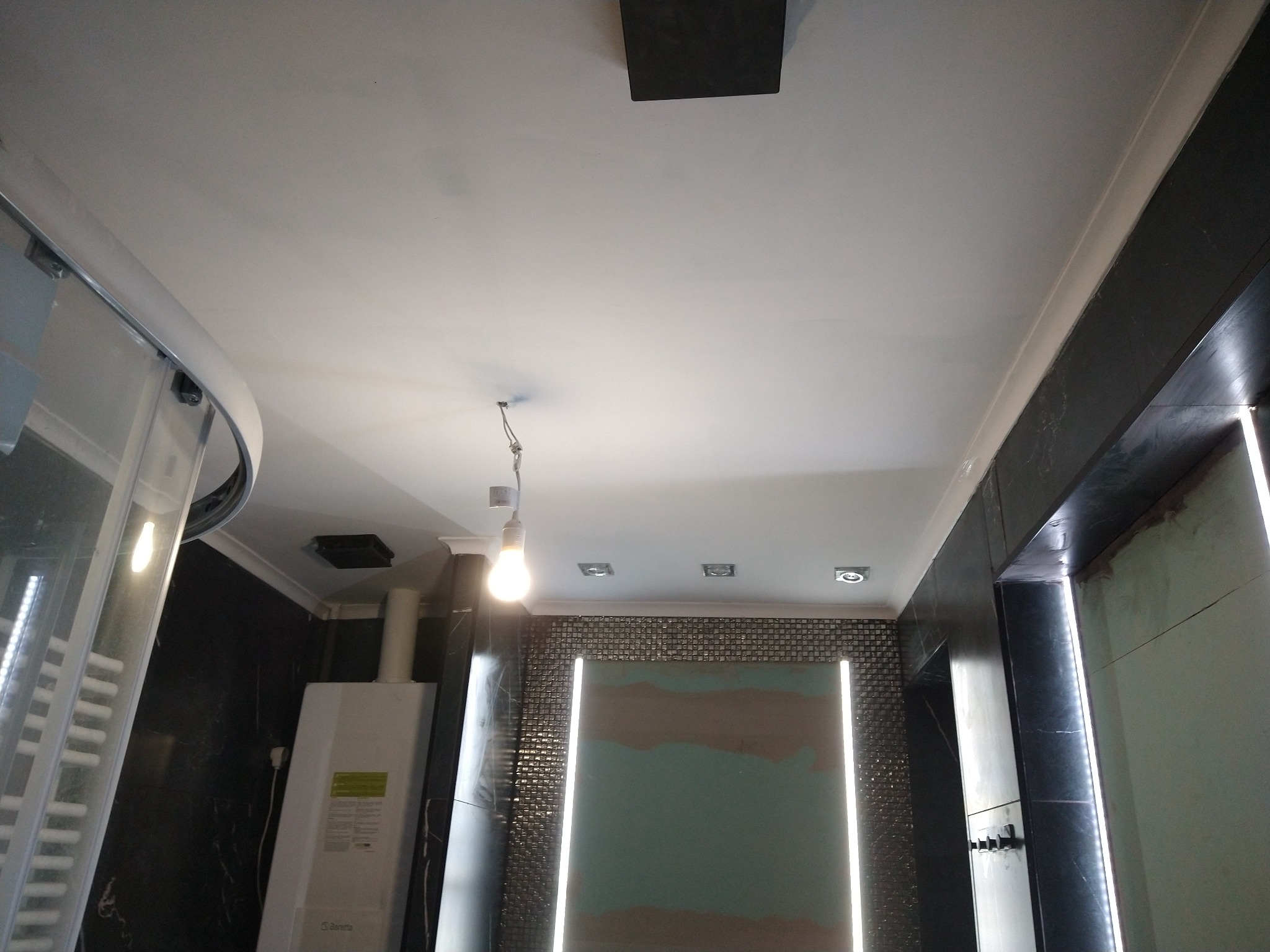 Łazienka w trakcie remontu: widoczna kabina prysznicowa, piec gazowy, ściana z mozaiki, podświetlane lustro i sufit z zamontowanym oświetleniem. Na ścianie widoczne ślady szpachlowania.