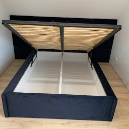 Zakład Stolarsko Budowlany HOLZ Rafał Bembnowicz - Otwarta rama łóżka z pojemnikiem na pościel, tapicerowana ciemnoniebieskim welurem, widoczne drewniane listwy stelaża i białe wnętrze pojemnika, jasne ściany w tle.