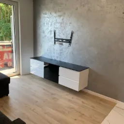 Nowoczesna szafka RTV wisząca, z białymi frontami i czarnym blatem, zamontowana na ścianie o dekoracyjnej fakturze, z widocznym uchwytem do telewizora i fragmentem minimalistycznego zegara.