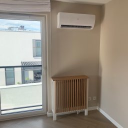 Zakład Stolarsko Budowlany HOLZ Rafał Bembnowicz - Biało-drewniana obudowa grzejnika z pionowymi listwami w jasnym pokoju z parkietem w jodełkę, klimatyzator na ścianie, widok z okna na balkon.