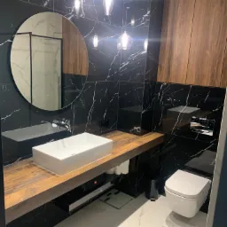 Nowoczesna łazienka w Miliczu z czarnymi marmurowymi płytkami, drewnianą szafką i blatem pod umywalkę, okrągłym lustrem oraz podwieszaną toaletą.