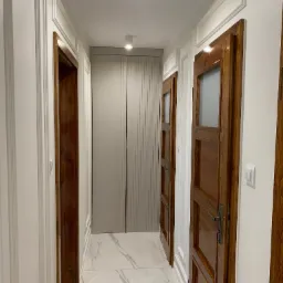 Wnętrze korytarza z dwiema drewnianymi drzwiami z przeszkleniami po bokach i szarą szafą na wprost, białe ściany z dekoracyjnymi listwami i marmurowa podłoga.