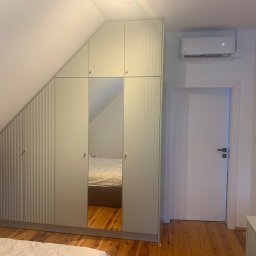 Zakład Stolarsko Budowlany HOLZ Rafał Bembnowicz - Jasna szafa wnękowa pod skosem dachu z lustrzanymi drzwiami, odbijającymi fragment sypialni, obok białe drzwi i klimatyzator.