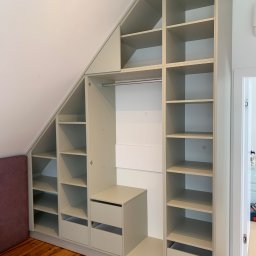 Zakład Stolarsko Budowlany HOLZ Rafał Bembnowicz - Zabudowa szaf wnękowych w szarym kolorze, dopasowana do skosu poddasza, z otwartymi półkami i szufladami, na drewnianej podłodze.