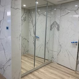 Zakład Stolarsko Budowlany HOLZ Rafał Bembnowicz - Nowoczesna szafa wnękowa z lustrzanymi drzwiami przesuwnymi i wykończeniem imitującym marmur, z widocznymi fragmentami wnętrza odbijającymi się w lustrze.