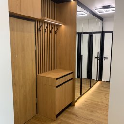 Zakład Stolarsko Budowlany HOLZ Rafał Bembnowicz - Nowoczesny przedpokój z zabudową z jasnego drewna: szafka na buty z szufladami, panel z wieszakami, szafka wisząca i lustro w wąskich ramach. Całość oświetlona listwami LED.