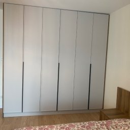 Zakład Stolarsko Budowlany HOLZ Rafał Bembnowicz - Biała szafa z sześcioma drzwiami z czarnymi uchwytami, wbudowana w ścianę sypialni, widoczna z perspektywy łóżka, z fragmentem szafki nocnej i wejściem do innego pomieszczenia z odkurzaczem w tle.