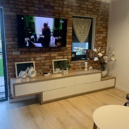 Zakład Stolarsko Budowlany HOLZ Rafał Bembnowicz - Salon z ceglaną ścianą, na której wisi telewizor i ozdobna serweta w oknie. Pod telewizorem biała szafka RTV z drewnianym blatem. Na blacie dekoracje, w tym białe ceramiczne wazony.