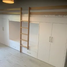 Biała antresola z drabinką i szafami po bokach, minimalistyczny design, drewniana drabinka, szare panele podłogowe, oświetlenie sufitowe.